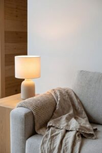 Close-up of a small minimalist white ceramic table lamp on a wooden side table next to a grey fabric sofa with a beige knit blanket and linen throw.